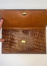 Brown leather clutch with crocodile texture and gold accents on a white background